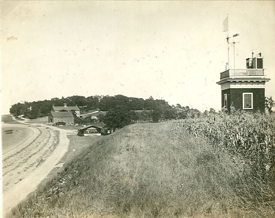 The image shows a vintage photograph of a coastal scene. A tall, dark tower with a white railing and a flag stands prominently on the right. A road winds along the coast on the left, leading towards a cluster of buildings nestled among trees. The foreground is dominated by a grassy hill.
